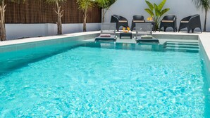 Piscina azul límpida com espreguiçadeiras e uma mesa, rodeada por um muro baixo e plantas.