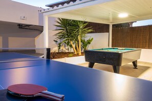 An outdoor pool table with a ping pong paddle and cue, a foosball table, and a potted plant.