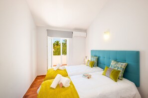A hotel room with two beds, a view of greenery outside, and an air conditioning unit on the wall.