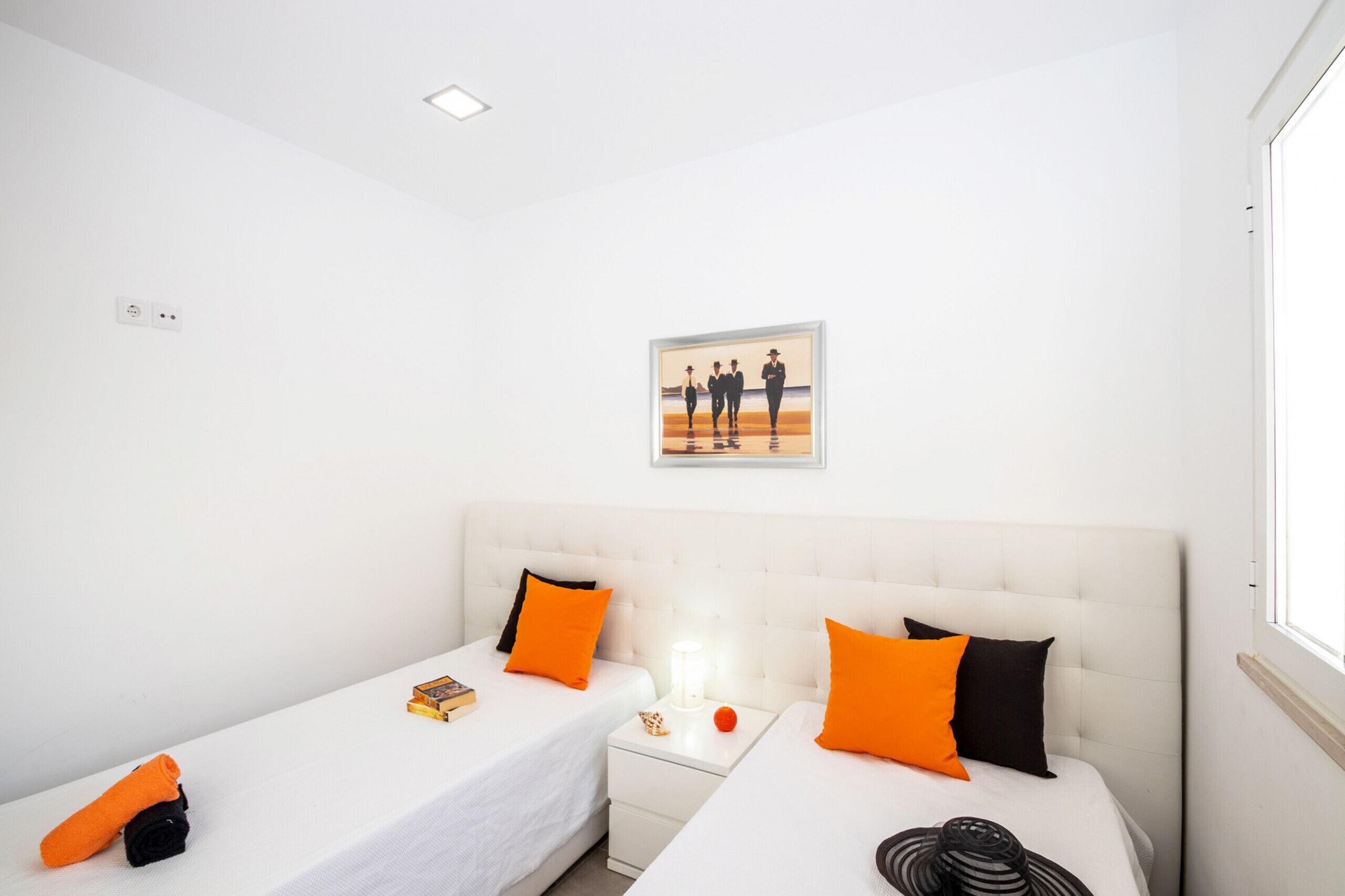 Ein Hotelzimmer mit zwei Betten, jeweils mit weißer Bettwäsche und orangenen Kissen, einem Nachttisch mit Lampe sowie einem gerahmten Bild an der Wand.