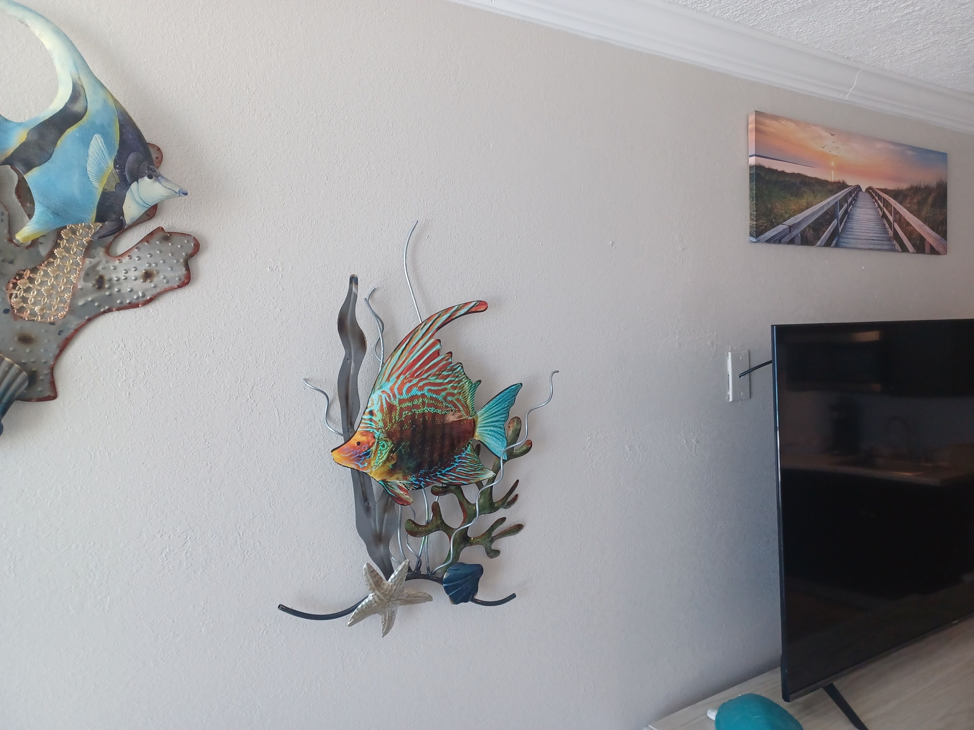 A wall with a decorative fish sculpture, a painting of a sunset, and a television.