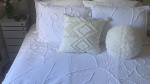 Una cama bien hecha con ropa de cama blanca y almohadas decorativas, una lámpara de noche y un florero con flores sobre una mesita de noche.