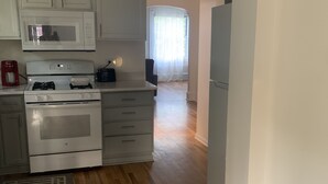 Una cocina con armarios blancos, microondas, estufa a gas y refrigerador. Hay una planta en la pared y una ventana con cortinas.