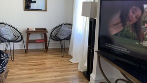 Sala de estar con televisor de pantalla plana, dos sillas negras, una mesita de madera, un espejo y una cortina blanca.