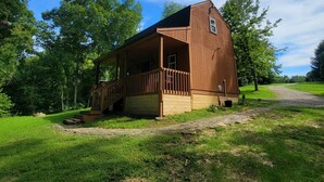 A wooden cabin with a porch, surrounded by lush greenery and a clear sky. - Rustic Private Cabin on 30 acres w/Loft/Firepit/Secluded/Trails (Patriot)