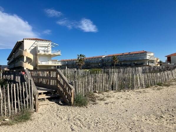 A beachfront property with a wooden fence, a staircase leading to a balcony, and a building with a red-tiled roof.