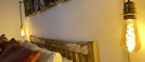 Una cama con cabecera rústica, una pared con tapiz floral y una bombilla colgante.