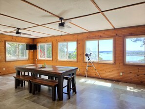 Une pièce aux murs lambrissés, comprenant une table à manger, des bancs, un télescope et de grandes fenêtres donnant sur l’extérieur.
