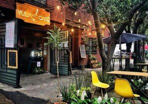 Un área de estar al aire libre con sillas y mesas amarillas, un edificio de ladrillo con un letrero que dice "YULA CAFE", y plantas en macetas.