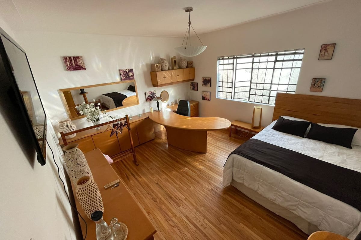 A modern bedroom with a bed, a desk, a chair, a vanity, and a large window.