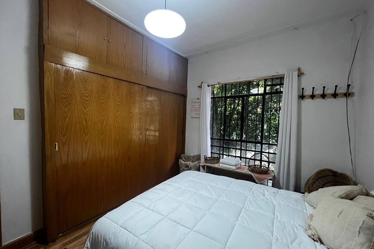 A bedroom with a wooden wardrobe, a bed with a white quilt, a window with a view of greenery, and a bedside table with a basket.