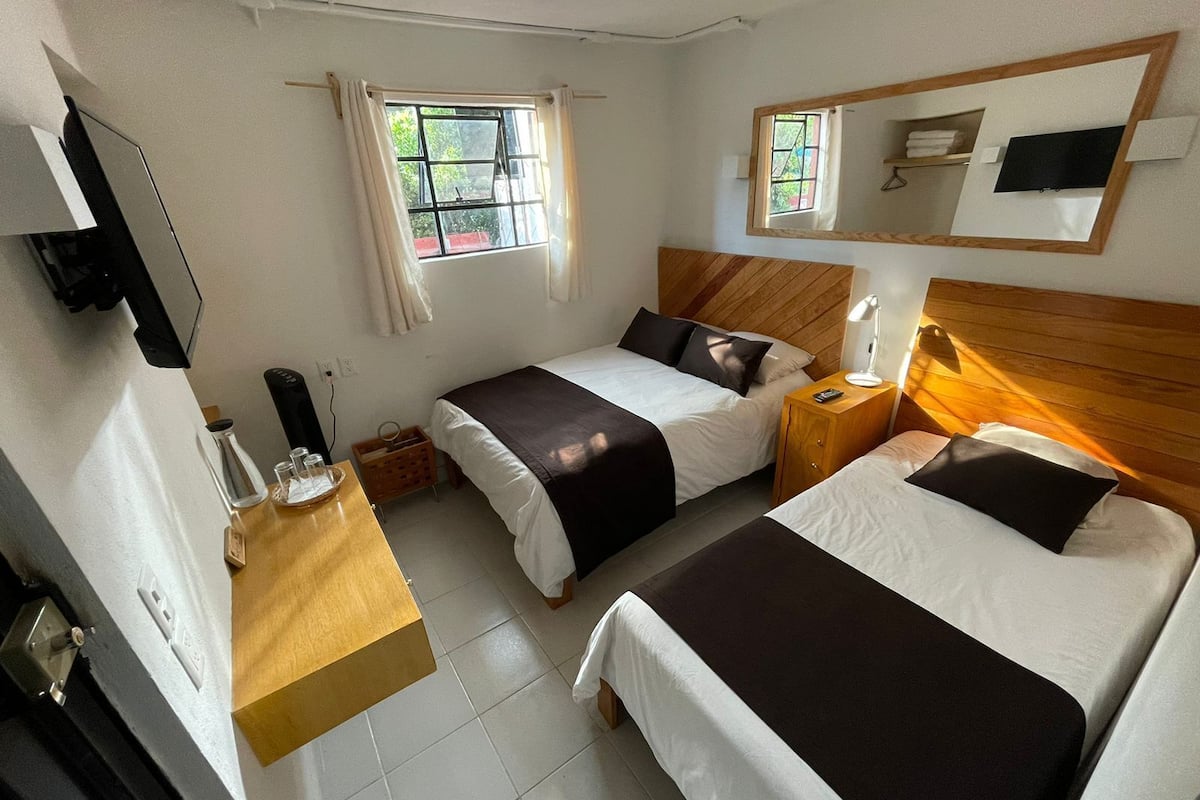 A hotel room with two beds, a wooden headboard, a bedside table with a lamp, a small desk with a lamp, and a window with curtains.