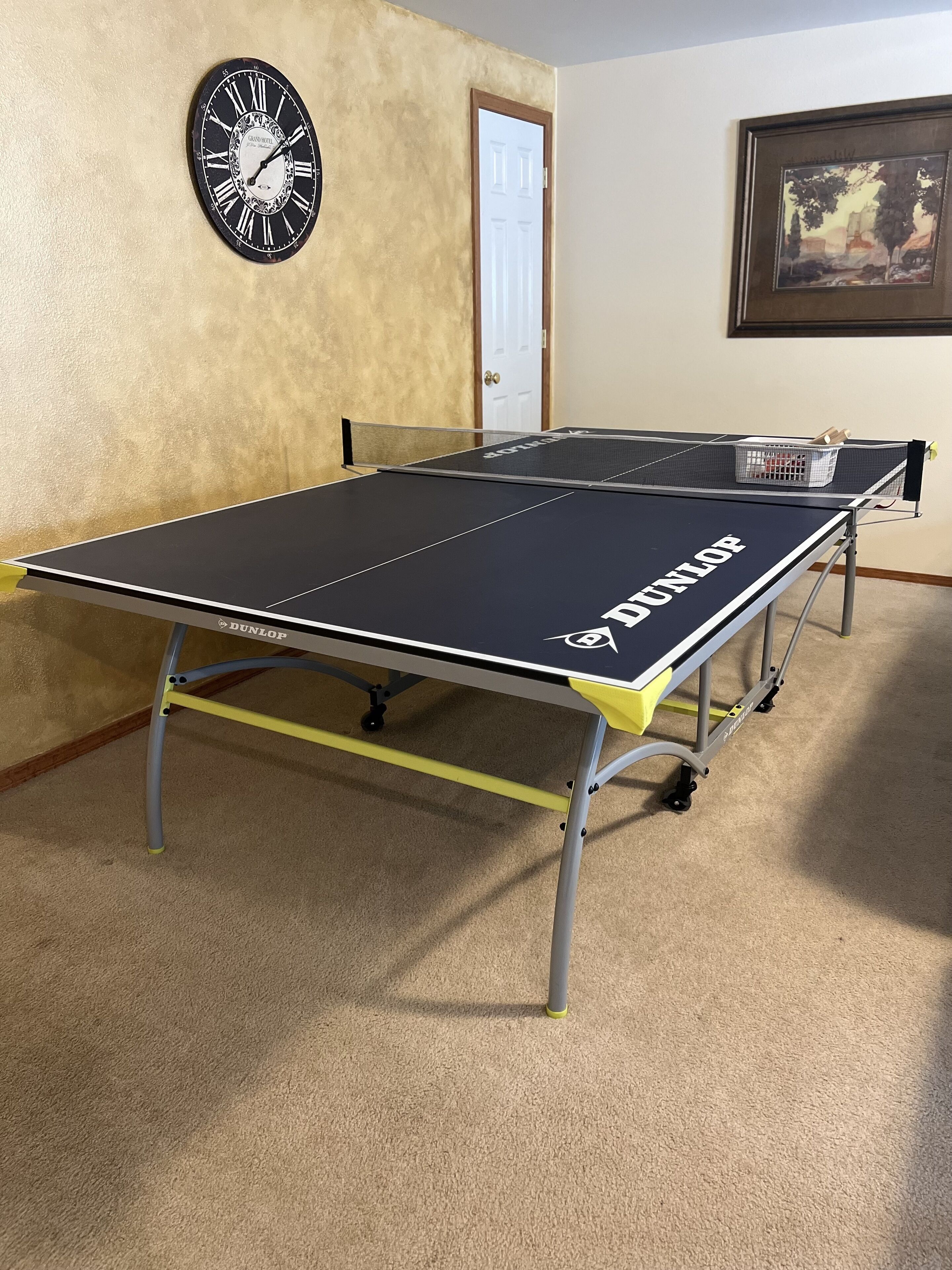 A ping pong table with a 'DUNLOP' logo, a wall clock, and a framed picture.