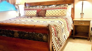 Uma cama de madeira com roupa de cama estampada, um criado-mudo com abajur e um quadro na parede.
