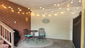 Ein überdachter Patio-Bereich mit Backsteinmauer, zwei Metallstühlen mit Kissen, einem kleinen Tisch und Lichterketten.