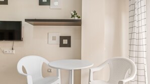 Una habitación con una mesa redonda blanca y dos sillas blancas, un televisor de pantalla plana montado en la pared, y una planta pequeña en maceta sobre una estantería.