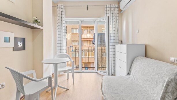 Habitación pequeña con cama, una mesita y una silla. Tiene balcón con vistas a otros edificios.