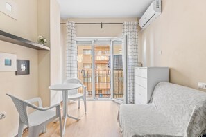 Une petite chambre comprenant un lit, une petite table et une chaise. Elle dispose d’un balcon avec vue sur d’autres immeubles.