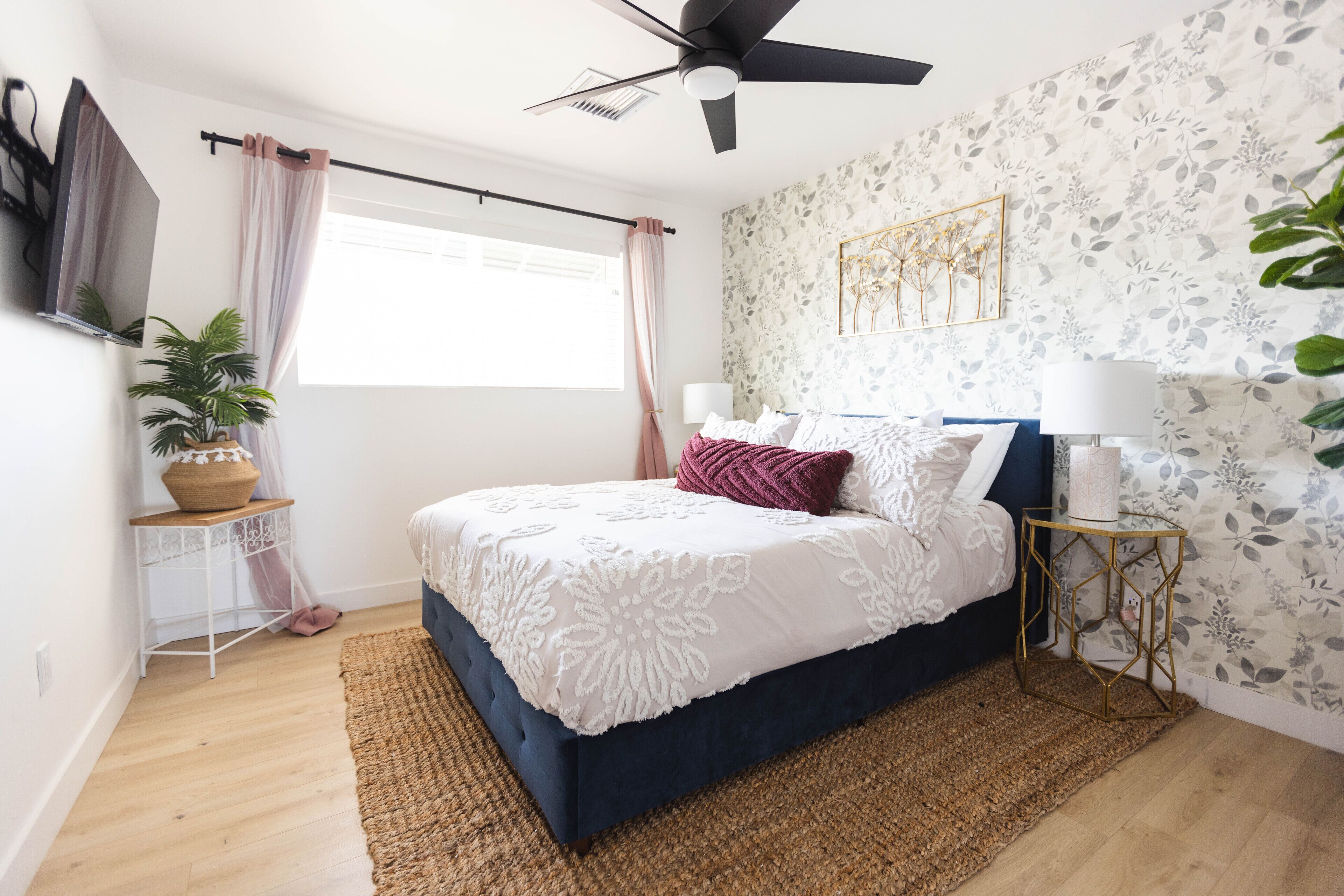 A bedroom with a bed, a ceiling fan, a window with curtains, a wall-mounted TV, a side table with a lamp, and a potted plant.
