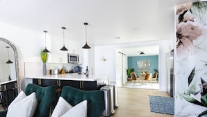 Un soggiorno moderno con un divano verde, una zona cucina con mobili bianchi e un'isola centrale, e un'area pranzo con una parete blu e un elemento decorativo a parete.