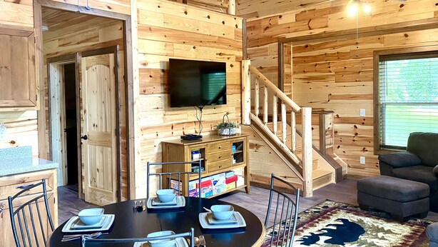 Een houten hut interieur met een eettafel gedekt voor vier personen, een aan de muur bevestigde televisie, en een trap naar een hoger niveau.