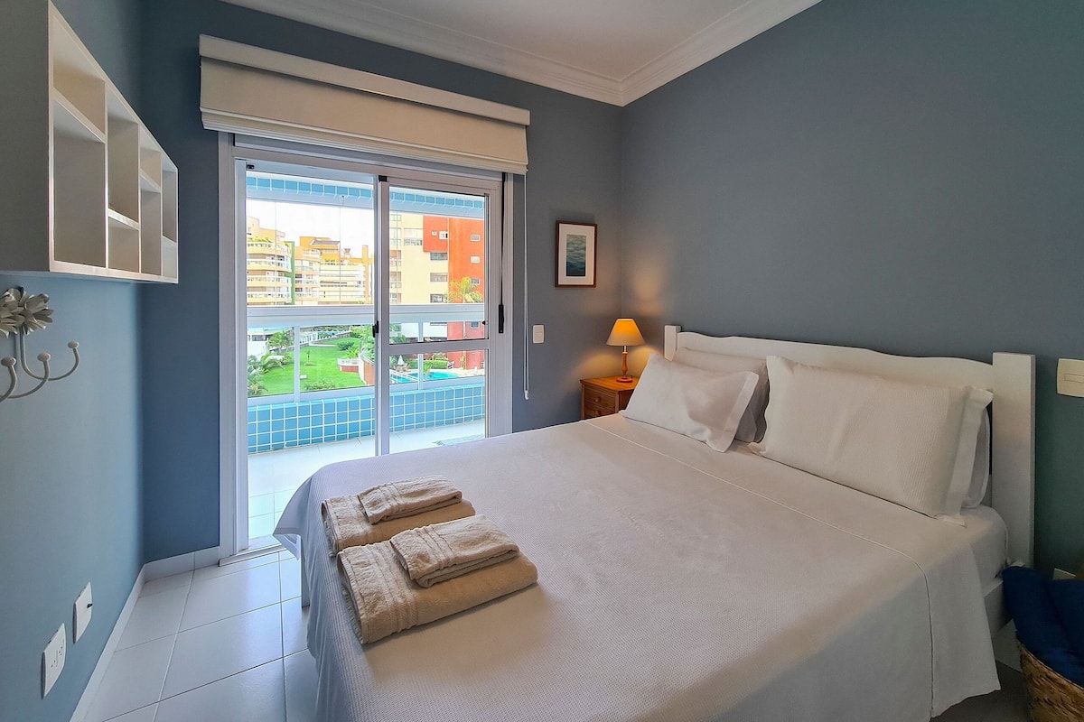 Un dormitorio con una cama grande, una ventana con vistas a edificios, una mesita de noche con una lámpara y un interruptor de luz en la pared.