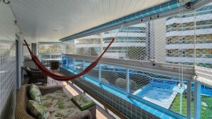 Un balcón techado con una hamaca, muebles de mimbre y vistas a una piscina y un edificio al fondo.