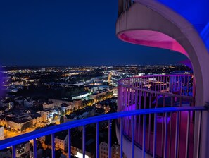 A nighttime cityscape viewed from a balcony with a railing.