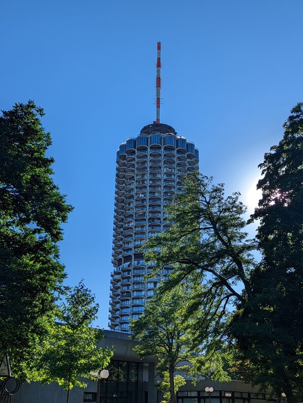 A tall, modern building with a distinctive antenna, surrounded by lush green trees.