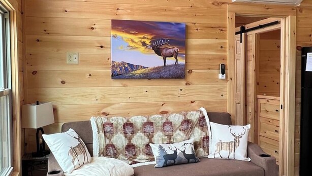 Interior acogedor de una cabaña con pared de madera, un cuadro de un ciervo, un sofá con cojines estampados y una mesita con una lámpara.