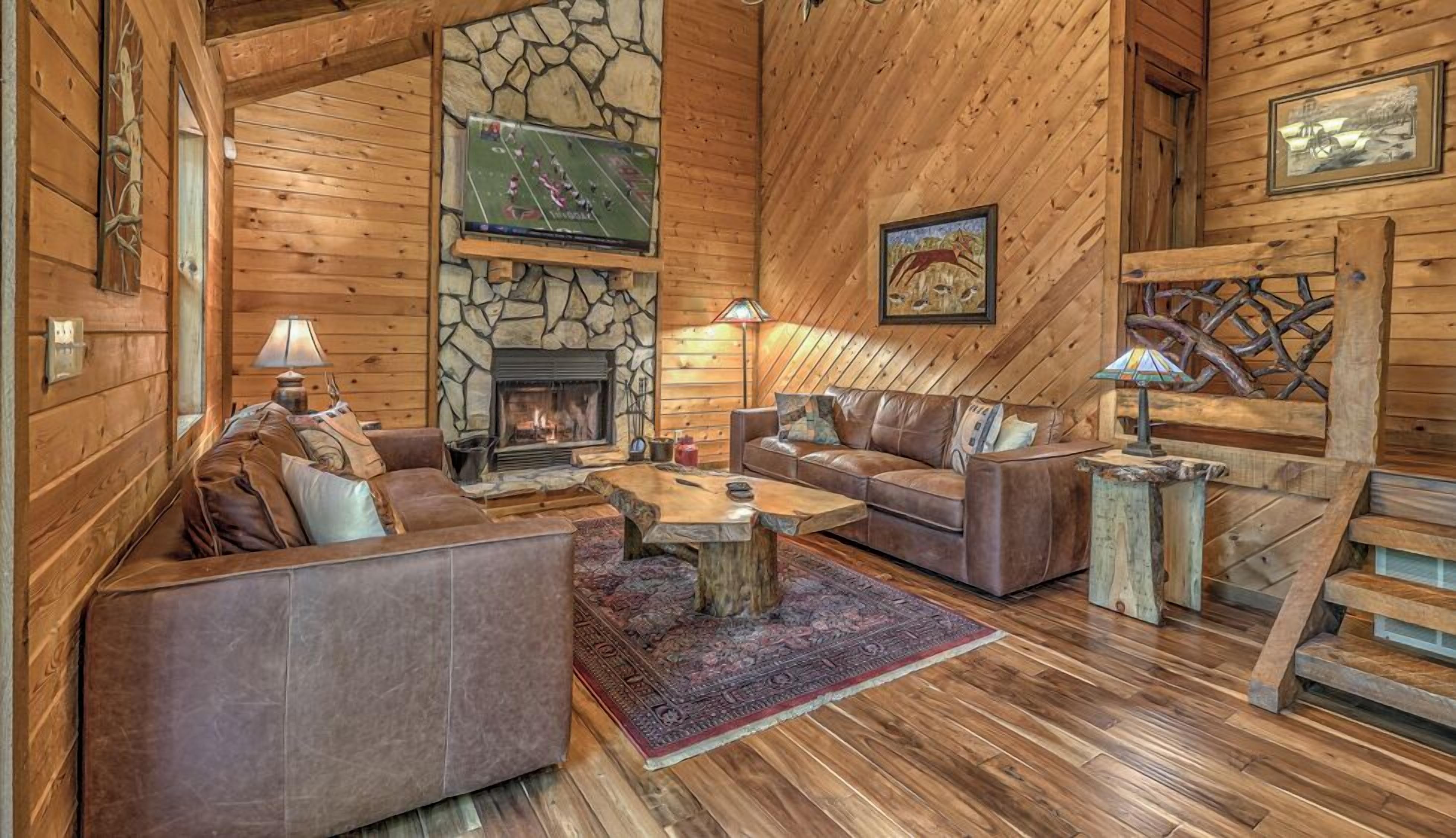 A cozy log cabin interior with a stone fireplace, leather sofas, and a rustic wooden floor.