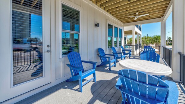 A covered outdoor patio with blue Adirondack chairs and a round table, wooden beams overhead, and large windows with blinds. - 🤗 E11G=Awesome! 6BR Beachview. Htd Pools. Easy beach access! (Gulf Shores)