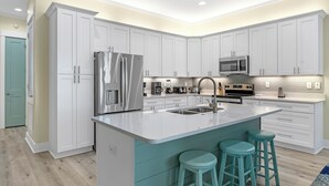 A modern kitchen with white cabinets, stainless steel appliances, a central island with stools, and a teal accent wall. - 🤗 E11G=Awesome! 6BR Beachview. Htd Pools. Easy beach access! (Gulf Shores)