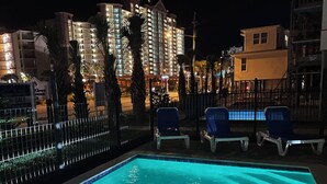A swimming pool with lounge chairs, surrounded by a fence, and buildings in the background. - 🤗 E11G=Awesome! 6BR Beachview. Htd Pools. Easy beach access! (Gulf Shores)