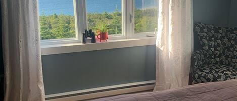 A bedroom with a large bed, a window with a view of the sea and greenery, and a potted plant on the windowsill.
