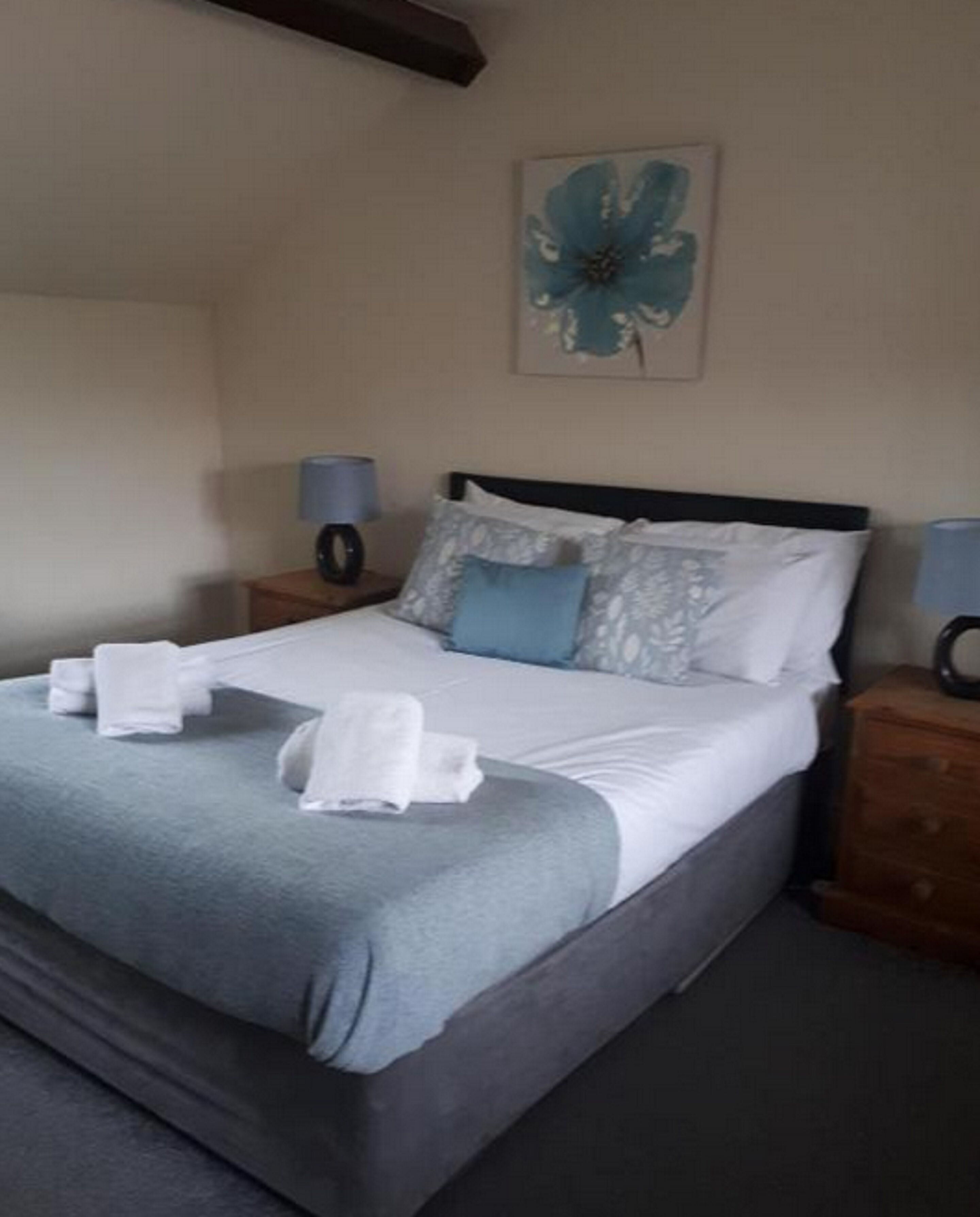 A neatly made bed with a grey and white duvet, two pillows, and a blue accent pillow. There are two bedside tables with lamps, and a framed artwork above the bed.