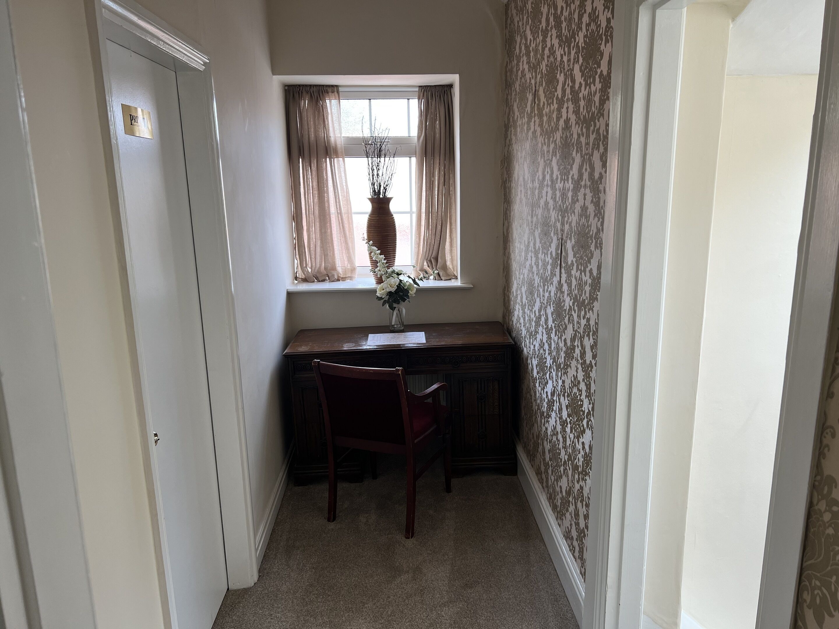 A narrow hallway with a desk and chair, a window with curtains, and a door labeled "PRV".