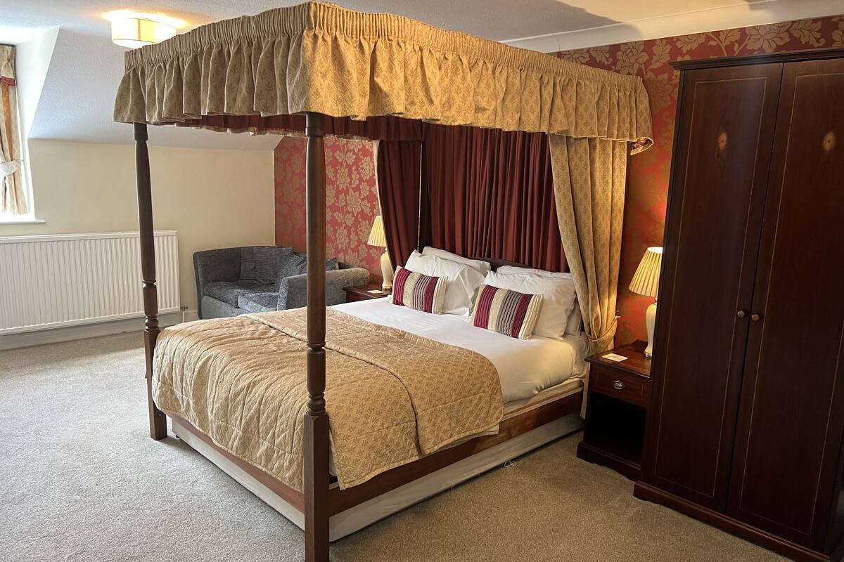 A four-poster bed with a canopy, a wooden wardrobe, and a sofa in a room with floral wallpaper.