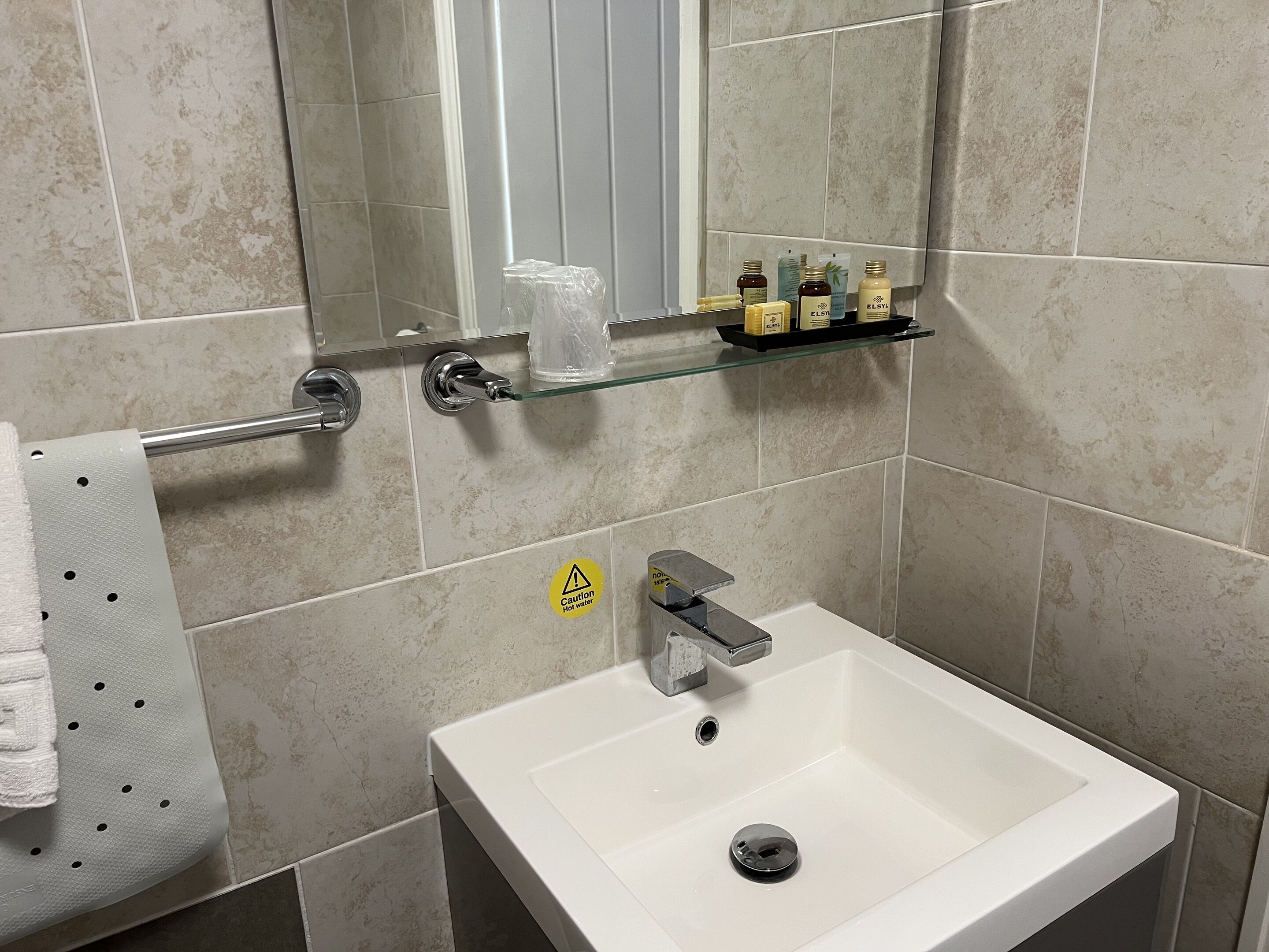 A bathroom with a white sink, a glass shelf with toiletries, a towel rack, and a caution sign for hot water.