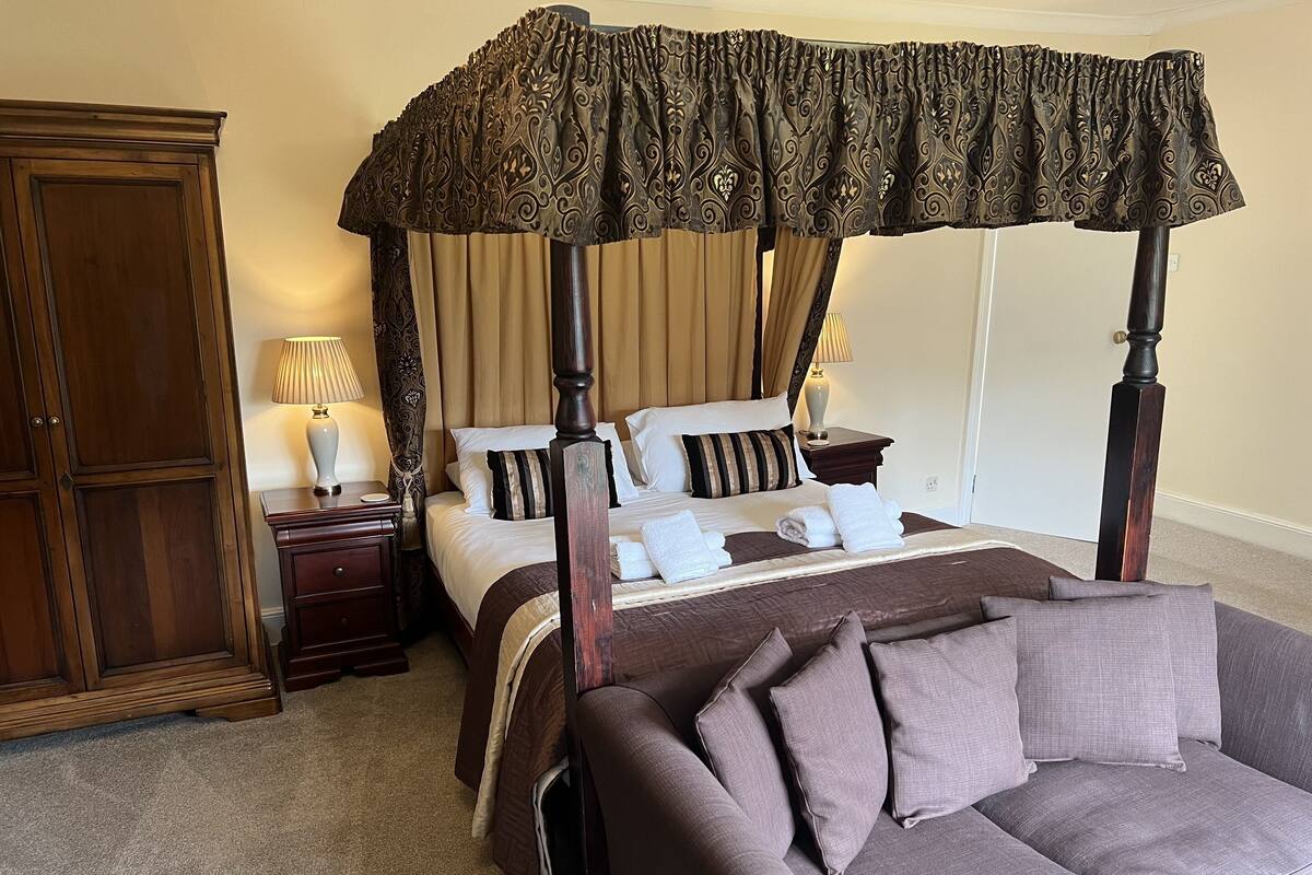 A four-poster canopy bed with a brown canopy, a wooden wardrobe, a nightstand with a lamp, and a grey sofa.