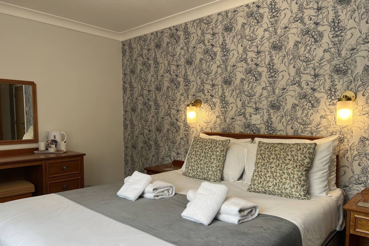 A neatly made bed with white and grey linens, a wooden nightstand with a lamp, and a floral wallpaper.