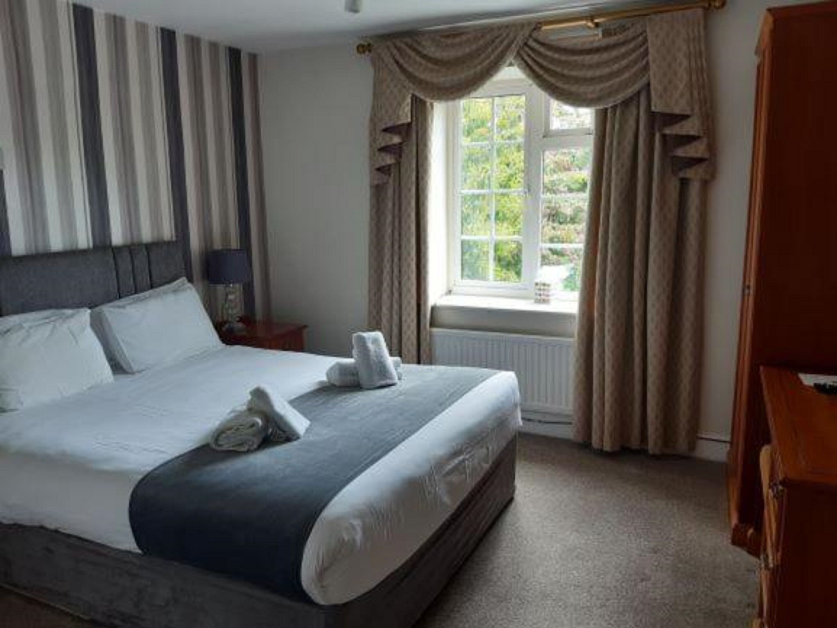 Een netjes opgemaakt bed met kussens en een opgevouwen handdoek, een nachtkastje met een lamp, een raam met gordijnen, en een radiator.