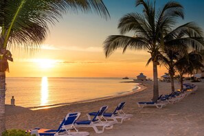 Una playa con filas de tumbonas azules y blancas, palmeras y una puesta de sol sobre el océano.
