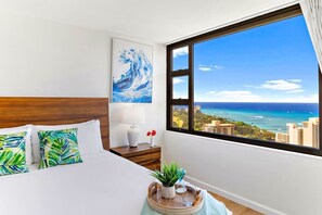Habitación de hotel con un ventanal grande, cama con sábanas blancas, cabecero de madera, mesita de noche con lámpara y vistas al mar y al paisaje urbano.