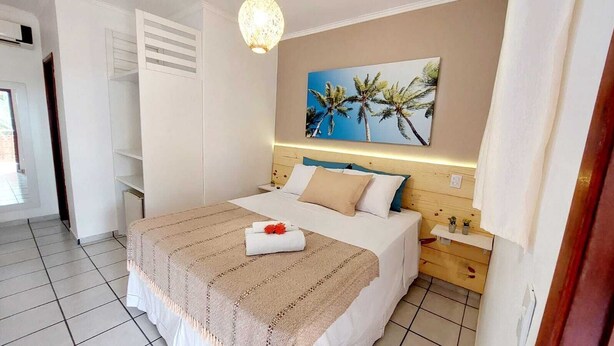 A hotel room with a bed, a nightstand, a wall-mounted lamp, and a painting of palm trees. - Pousada Anauê (Conde)