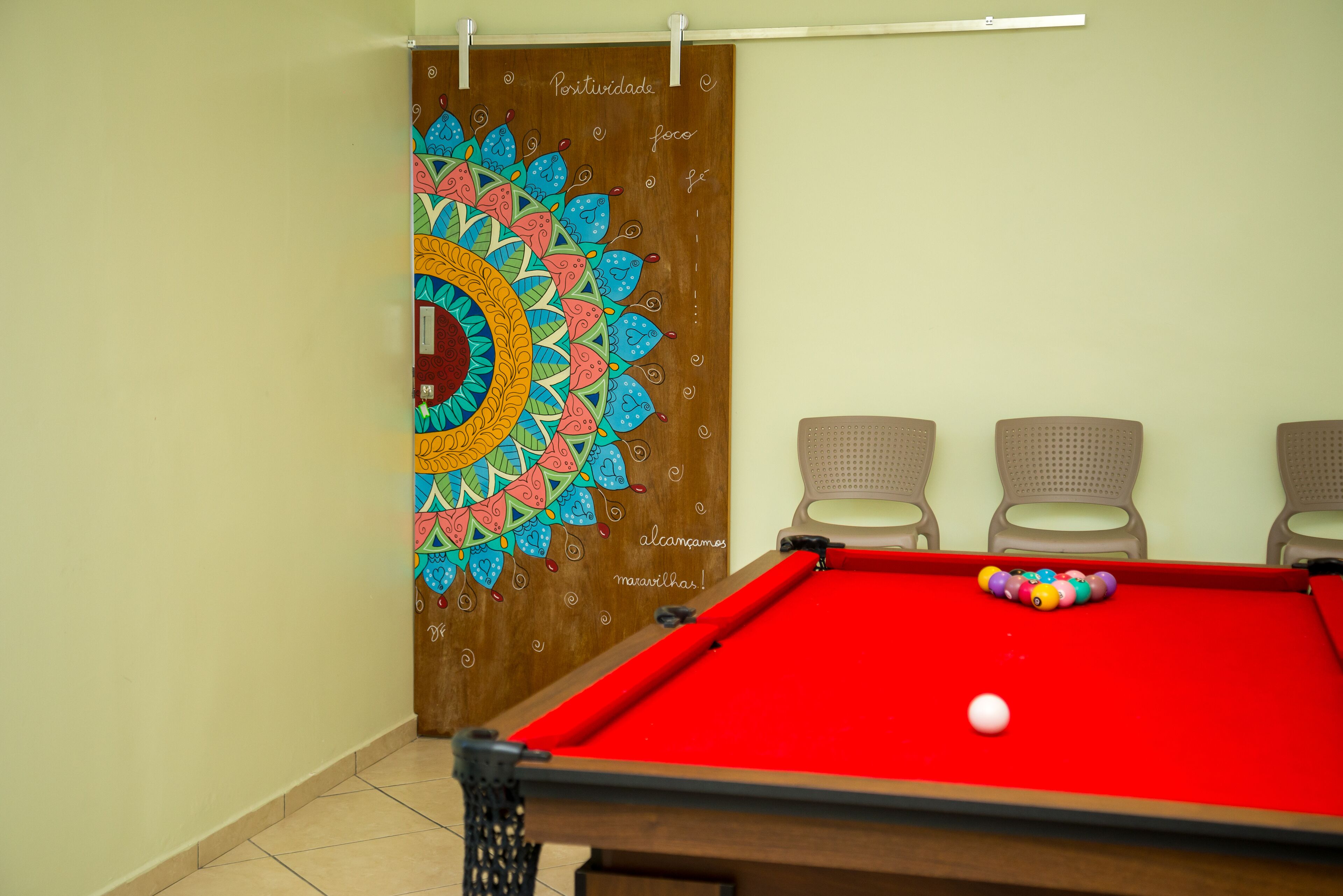 A pool table with a red felt surface, a colorful mural on the wall, and three beige chairs.