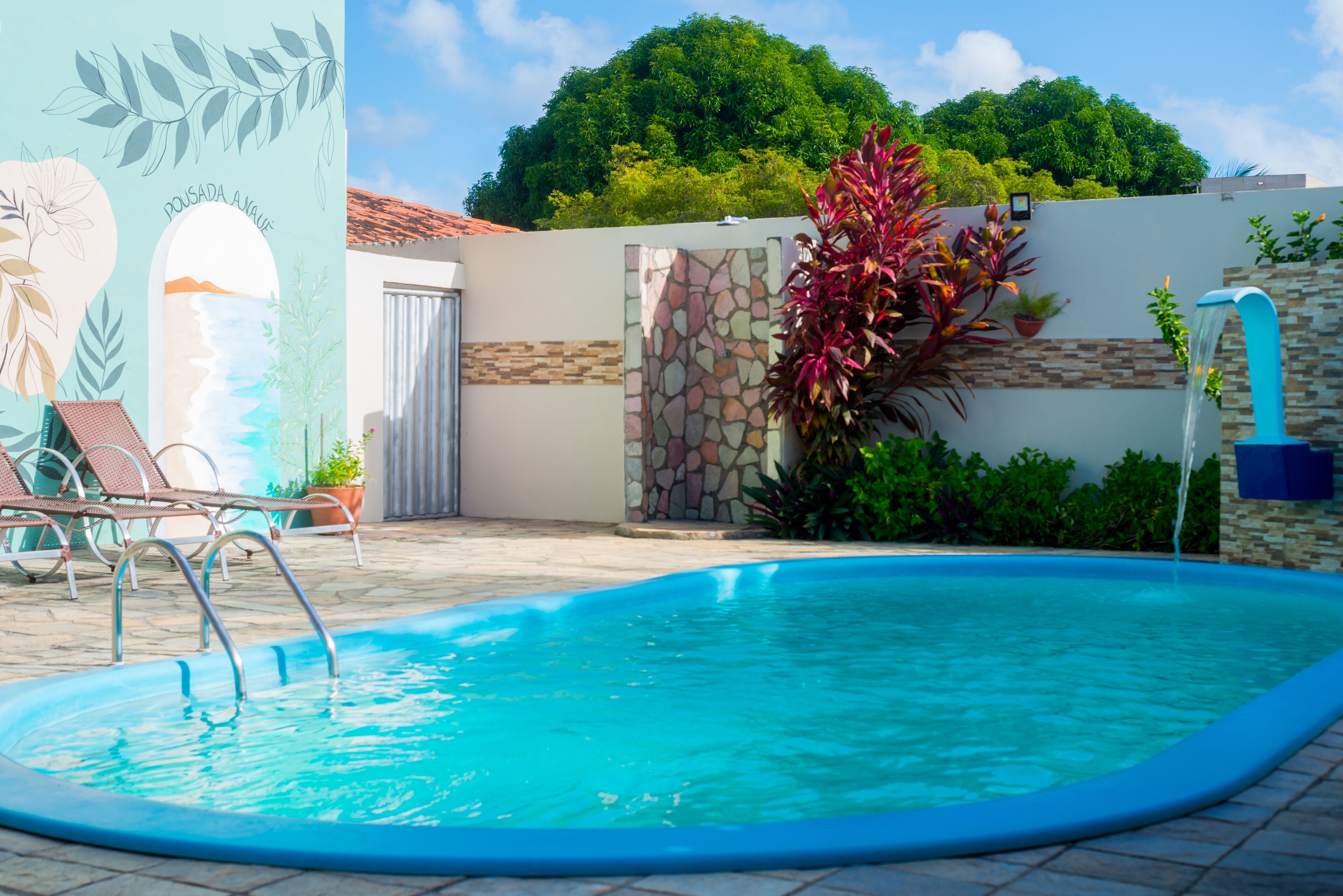 Una piscina con un muro de piedra y un mural, rodeada de sillones de descanso y plantas en macetas.