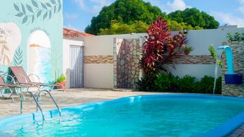 Una piscina con un muro de piedra y un mural, rodeada de sillones de descanso y plantas en macetas.
