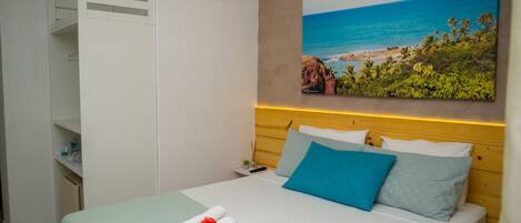 Una cama bien hecha con una colcha verde, una almohada azul y una almohada blanca. En la pared sobre la cama, hay un cuadro de una escena de playa.