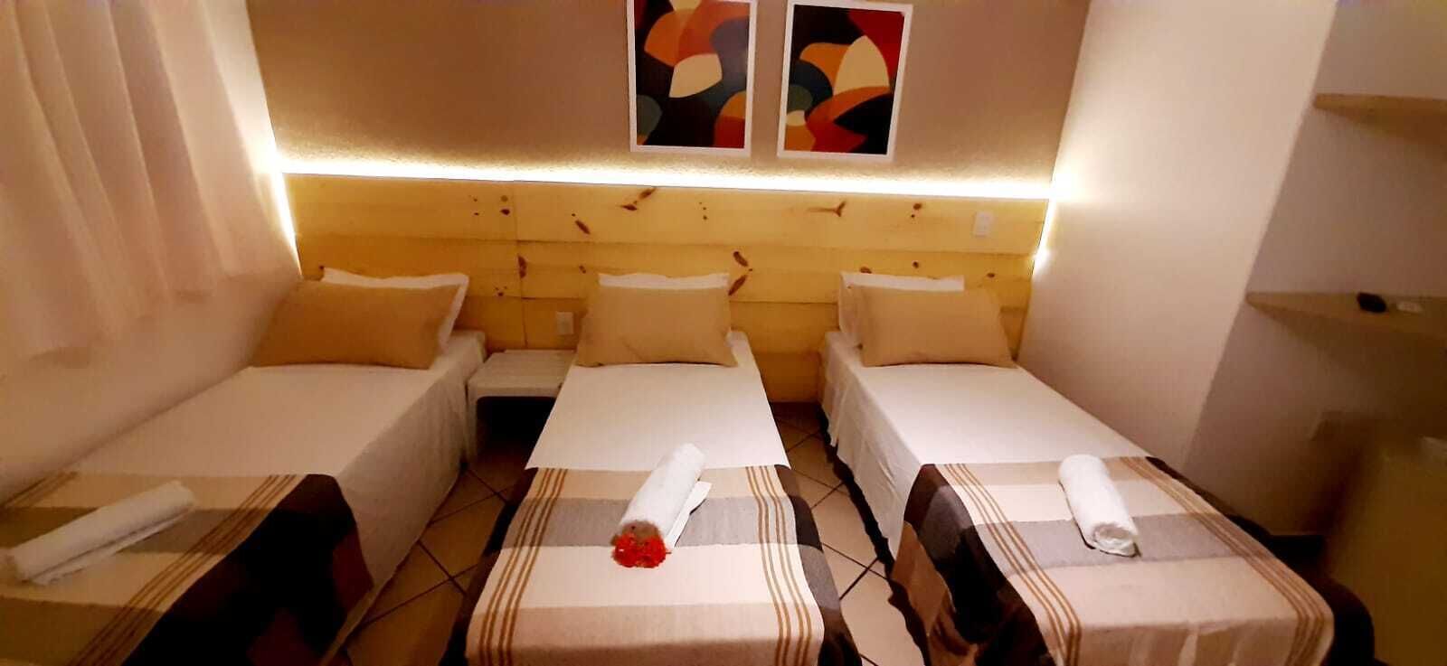 A hotel room with three single beds, each with a pillow and a rolled towel. There are two abstract paintings on the wall above the beds.
